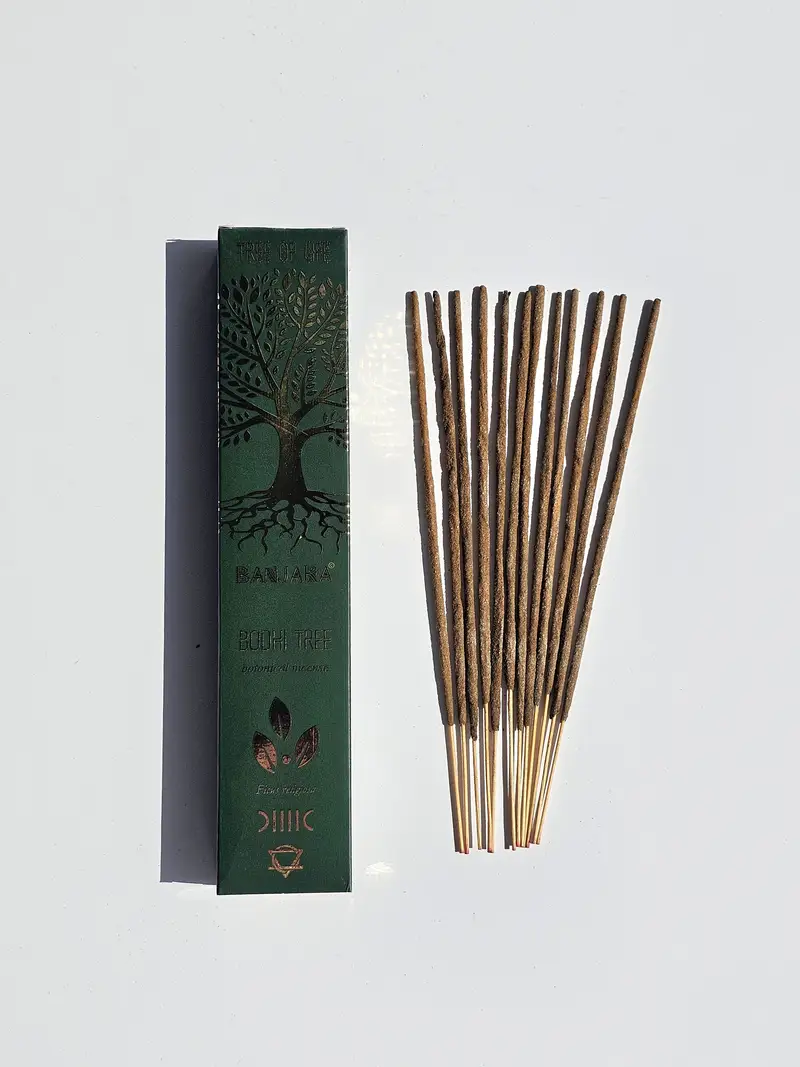 Ciemnozielone pudełko kadzidełek Banjara Bodhi Tree obok grubych naturalnych patyczków botanicznych i tlącego się kadzidła na podstawce ceramicznej.
