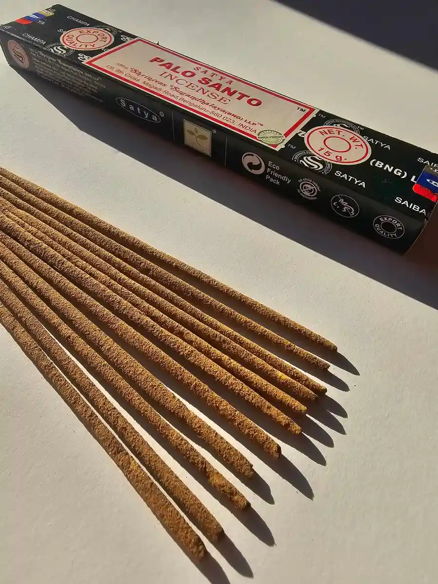 Kadzidła Satya White Sage 15g w jasnoszarym kartonowym pudełku, pod którym leży kilkanaście patyczków z jasnobrązową masalą na białym tle z mocnym cieniem.