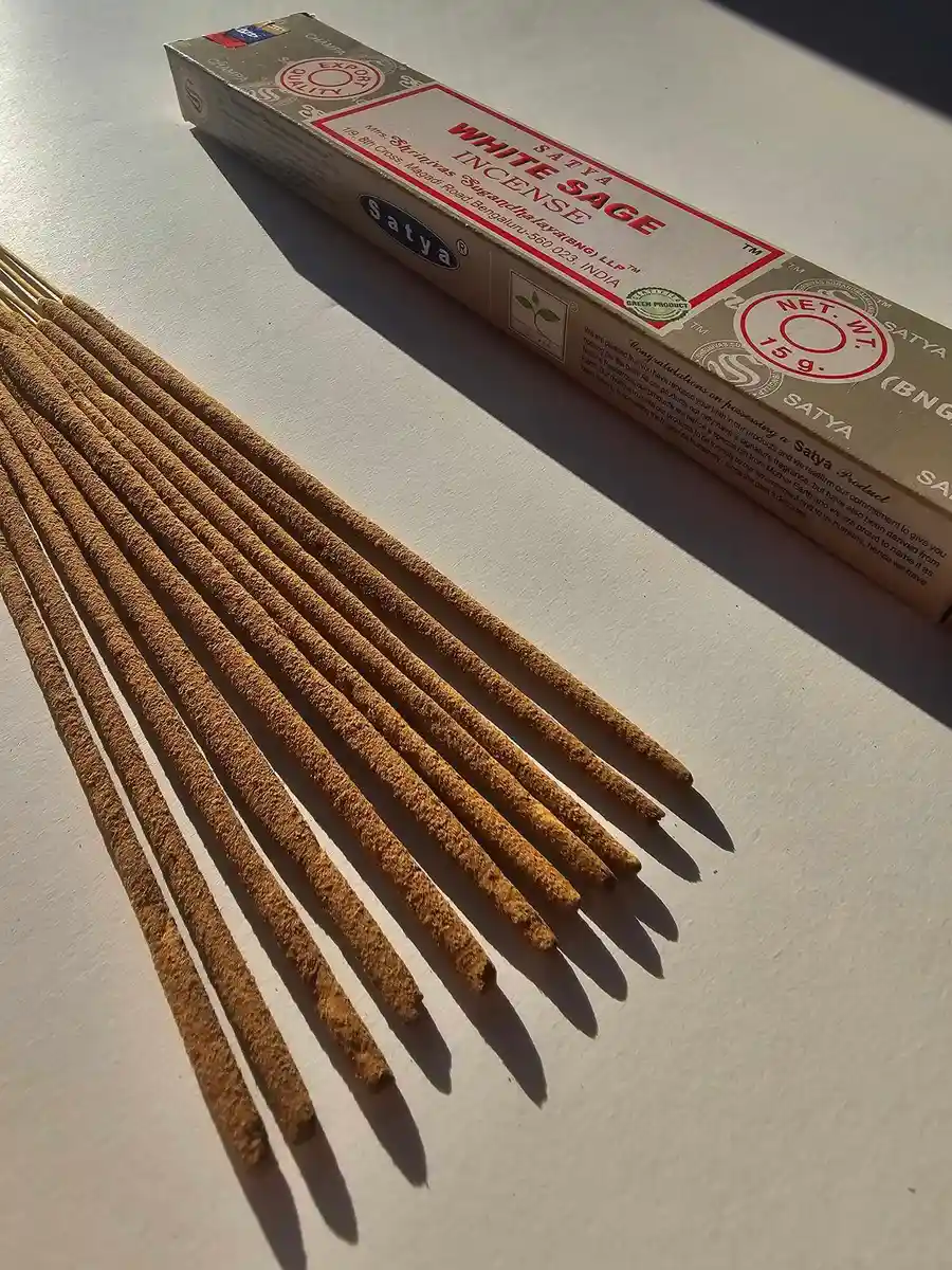 Kadzidła Satya White Sage 15g w beżowym, papierowym pudełku z logo marki, obok wysypane patyczki kadzidlane na szarym tle z głębokimi cieniami.