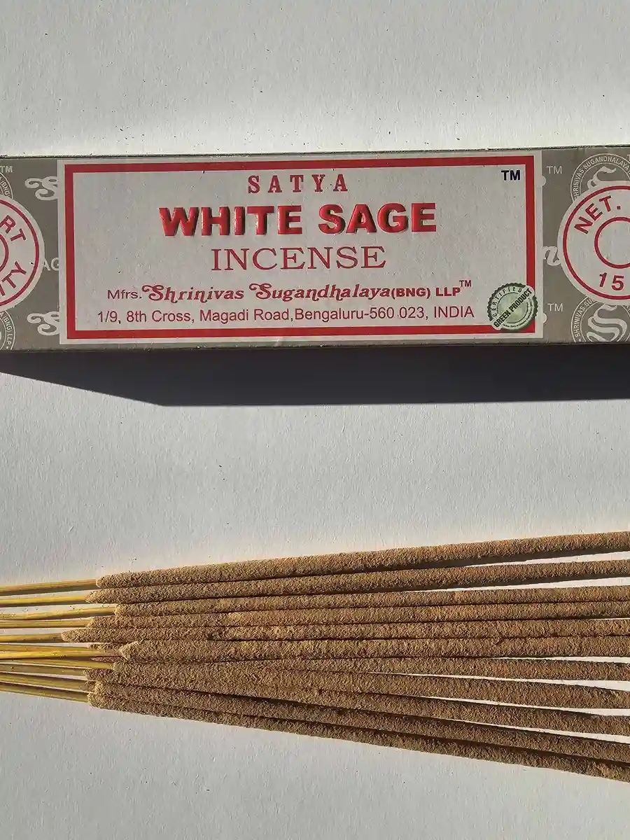 Kadzidła Satya White Sage 15g w beżowym, papierowym pudełku z logo marki, obok wysypane patyczki kadzidlane na szarym tle z głębokimi cieniami.