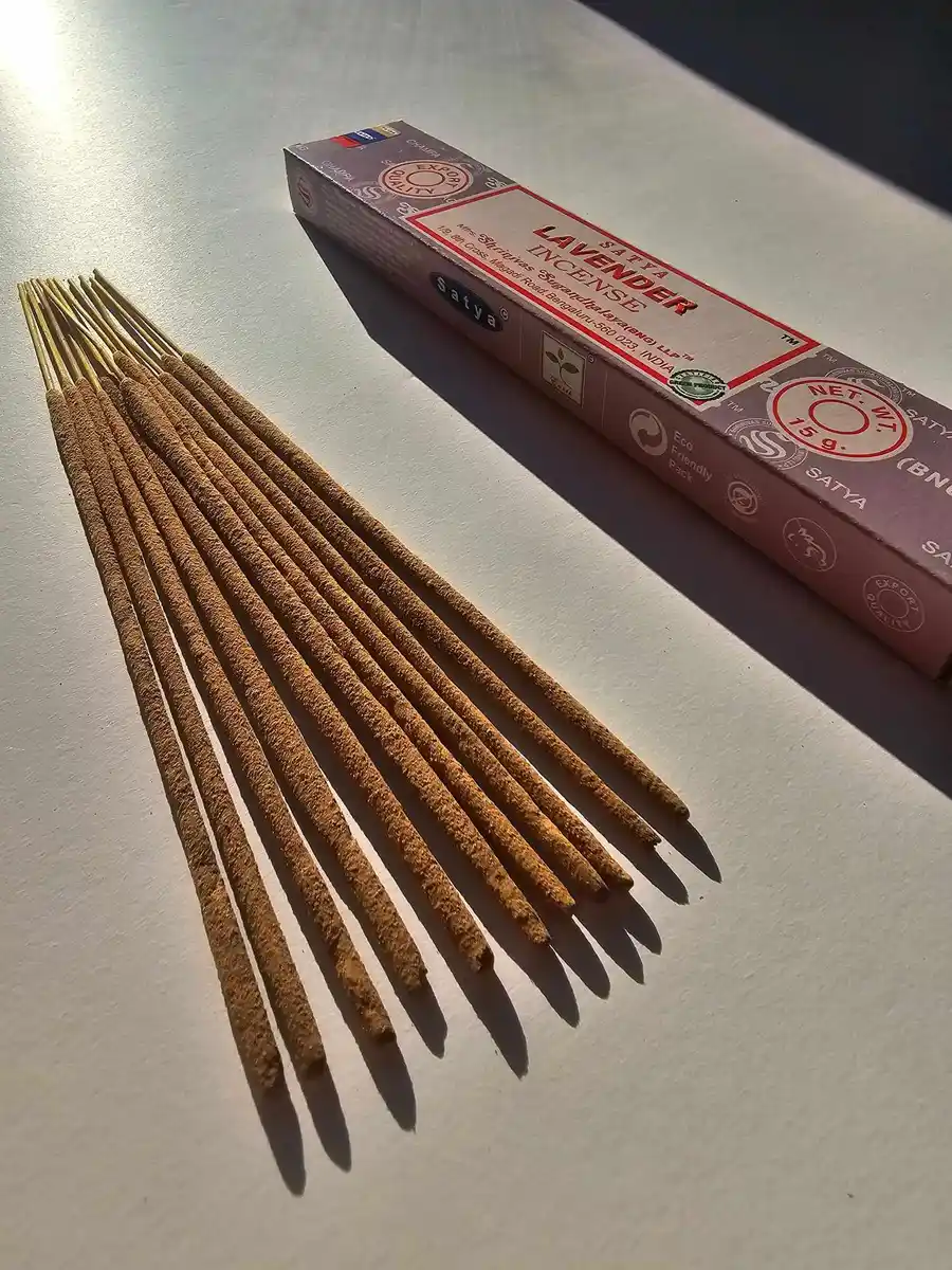 Jasnofioletowe pudełko kadzideł Satya Lavender 15g leżące nad stosem jasnobrązowych patyczków na białym tle z wyraźnym cieniem.