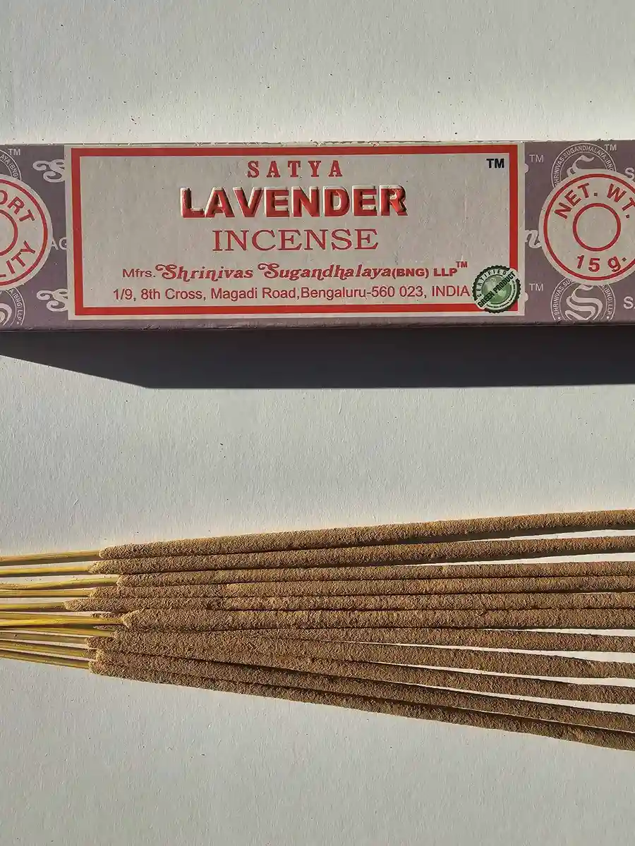 Jasnofioletowe pudełko kadzideł Satya Lavender 15g leżące nad stosem jasnobrązowych patyczków na białym tle z wyraźnym cieniem.