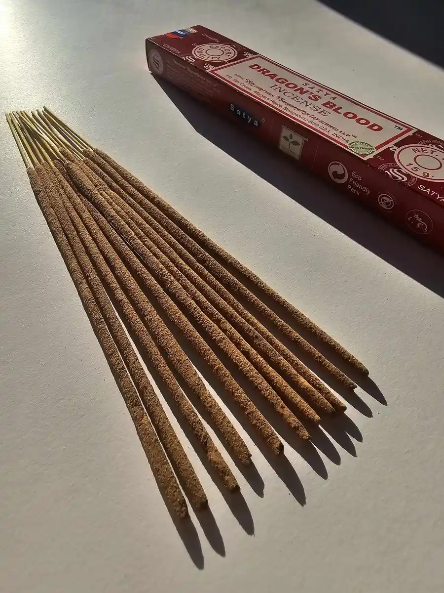 Czerwone pudełko kadzideł Satya Dragon's Blood 15g leżące nad grupą jasnobrązowych patyczków na białym tle z wyraźnym, ostrym cieniem.