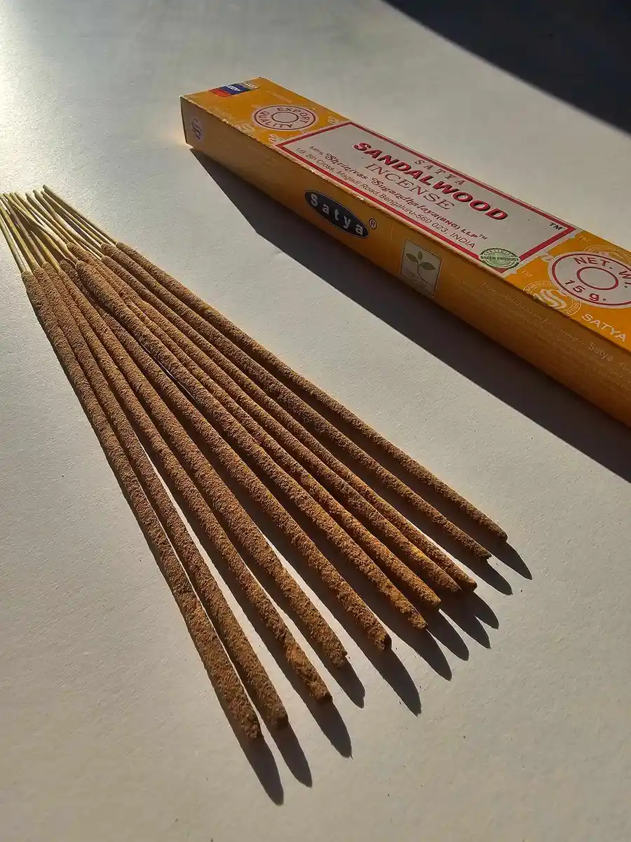 Żółto-pomarańczowe pudełko kadzideł Satya Sandalwood 15g leżące nad grupą jasnobrązowych patyczków na białym tle z wyraźnym poziomym cieniem.
