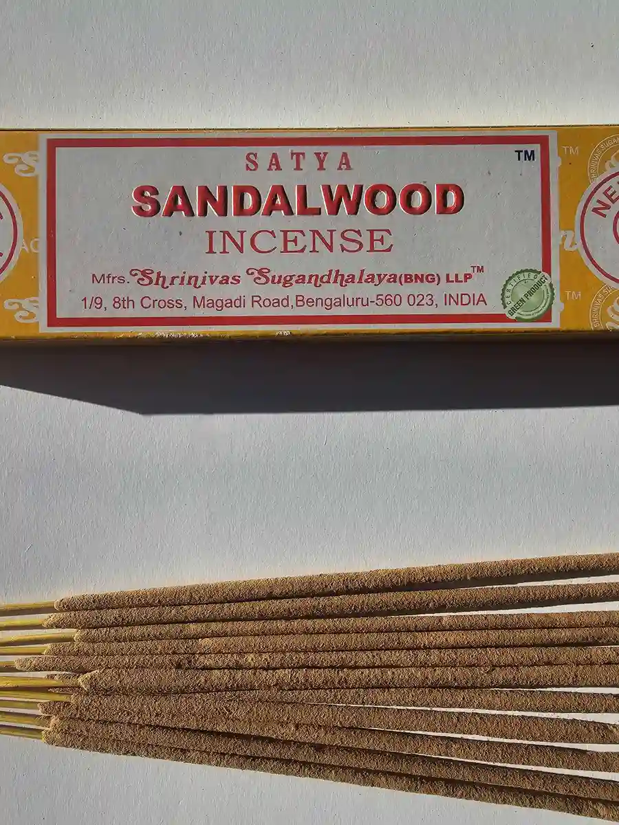 Żółto-pomarańczowe pudełko kadzideł Satya Sandalwood 15g leżące nad grupą jasnobrązowych patyczków na białym tle z wyraźnym poziomym cieniem.