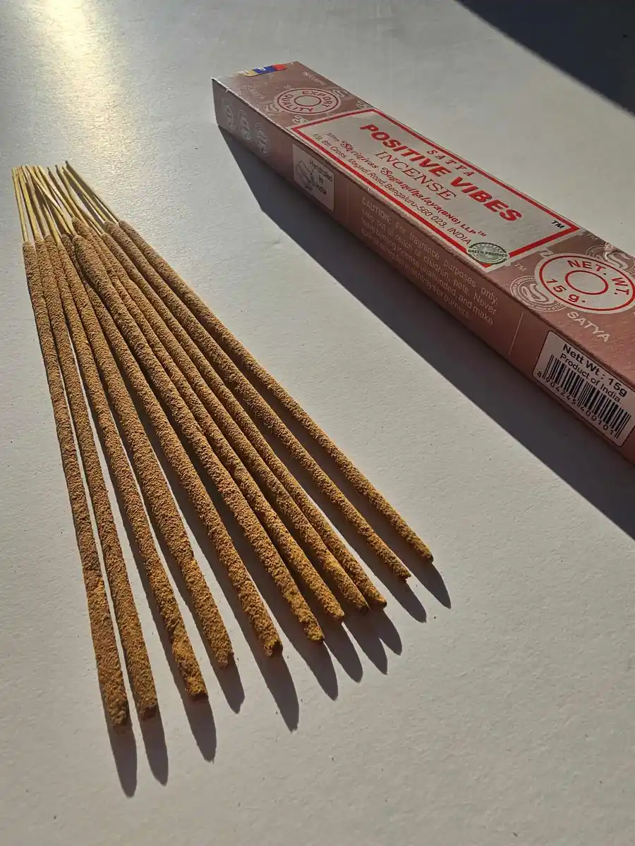 Jasnobrązowe pudełko kadzideł Satya Positive Vibes 15g z czerwonymi napisami, ułożone nad stosem jasnobrązowych patyczków na białym tle z cieniem.