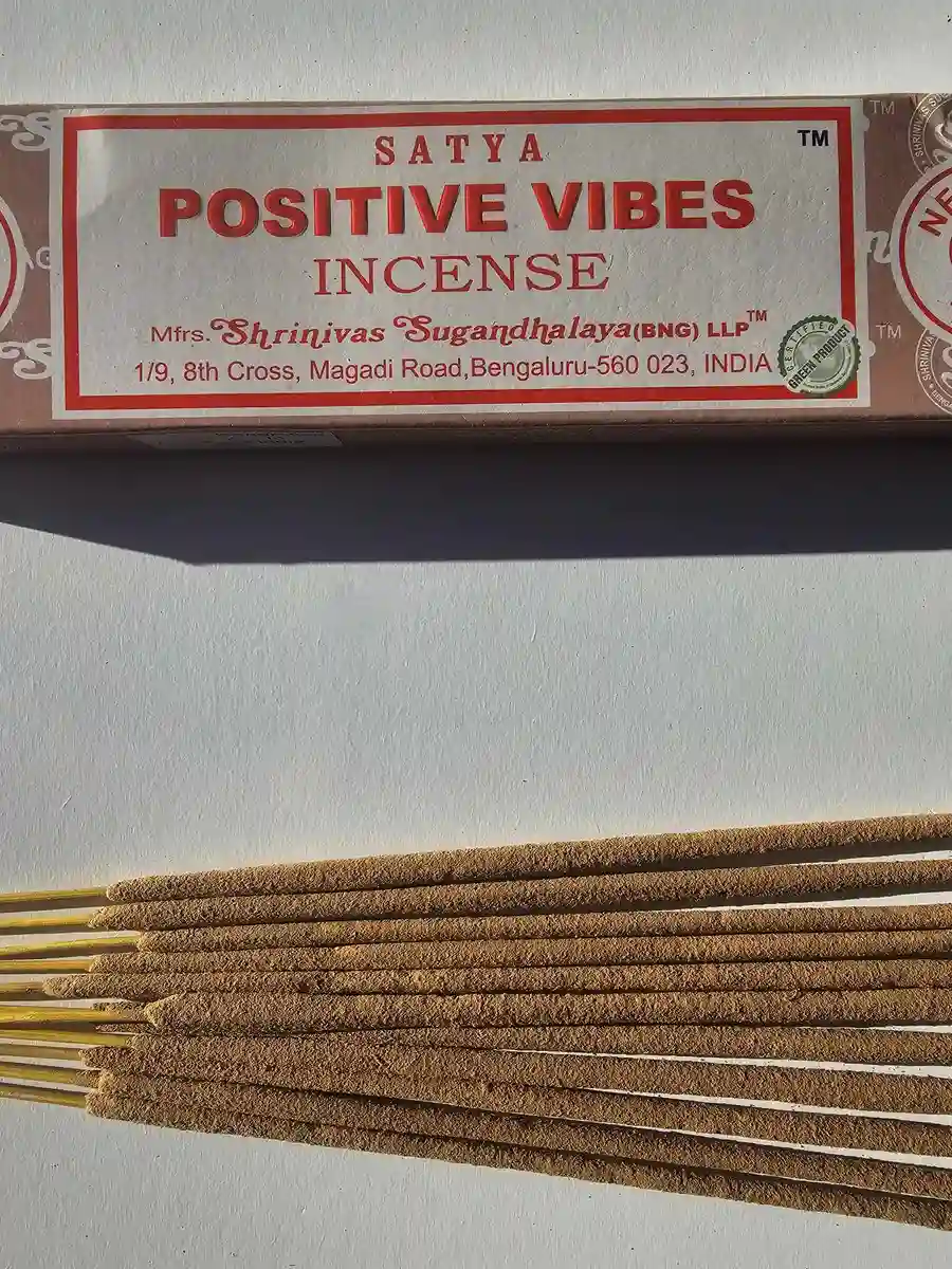 Jasnobrązowe pudełko kadzideł Satya Positive Vibes 15g z czerwonymi napisami, ułożone nad stosem jasnobrązowych patyczków na białym tle z cieniem.