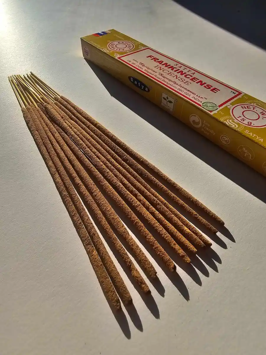 Żółte pudełko kadzideł Satya Frankincense 15g leżące nad stosem jasnobrązowych patyczków na białym tle z wyraźnym, poziomym cieniem.