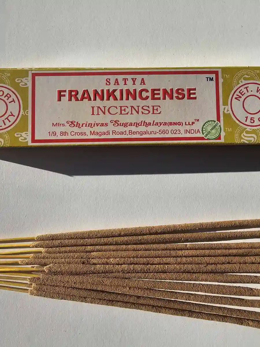 Żółte pudełko kadzideł Satya Frankincense 15g leżące nad stosem jasnobrązowych patyczków na białym tle z wyraźnym, poziomym cieniem.