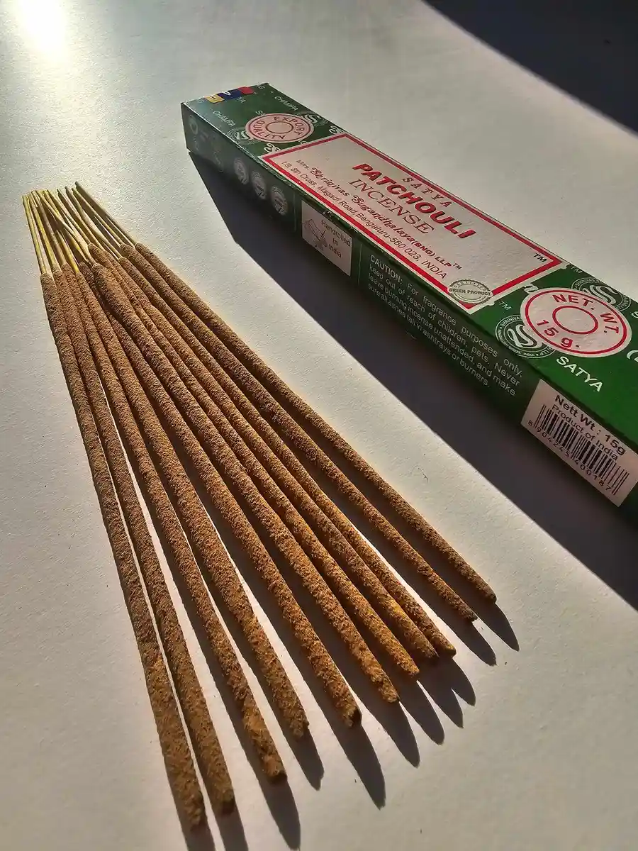 Zielone pudełko kadzideł Satya Patchouli 15g leżące nad grupą jasnobrązowych patyczków na białym tle z wyraźnym, poziomym cieniem.