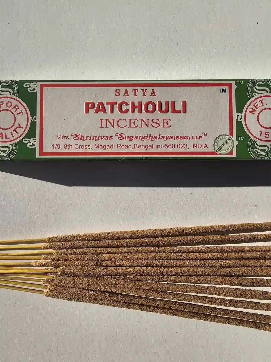 Zielone pudełko kadzideł Satya Patchouli 15g leżące nad grupą jasnobrązowych patyczków na białym tle z wyraźnym, poziomym cieniem.