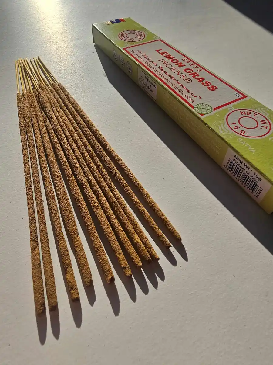 Jasnozielone pudełko kadzideł Satya Lemon Grass 15g ułożone nad stosem jasnobrązowych patyczków na białym tle z wyraźnym cieniem.
