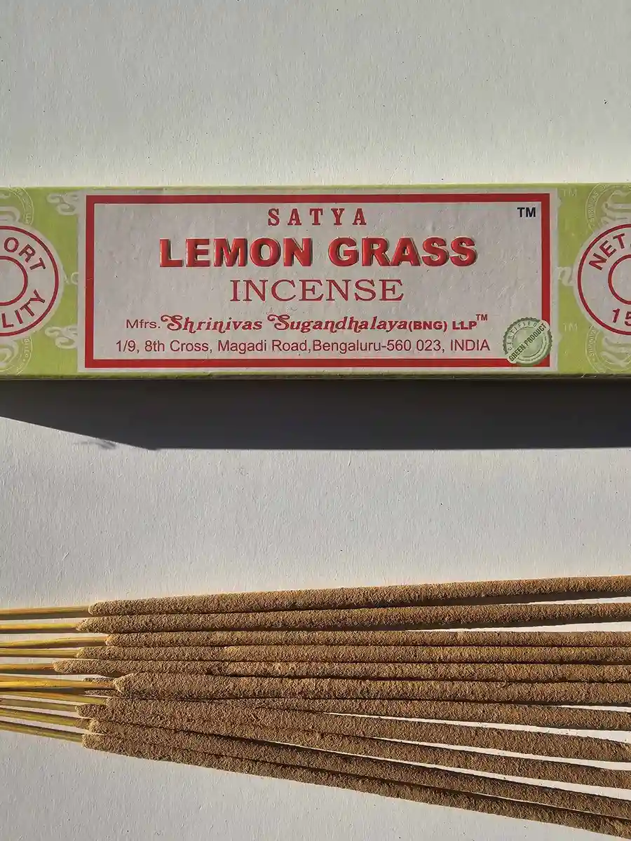 Jasnozielone pudełko kadzideł Satya Lemon Grass 15g ułożone nad stosem jasnobrązowych patyczków na białym tle z wyraźnym cieniem.