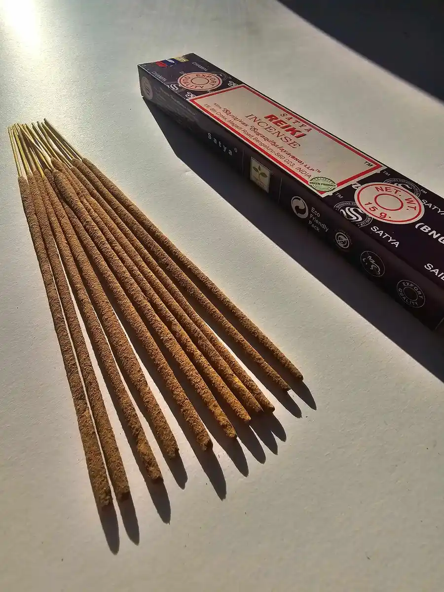 Ciemnofioletowe pudełko kadzideł Satya Reiki 15g umieszczone nad stosem jasnobrązowych patyczków na białym tle z wyraźnym cieniem.