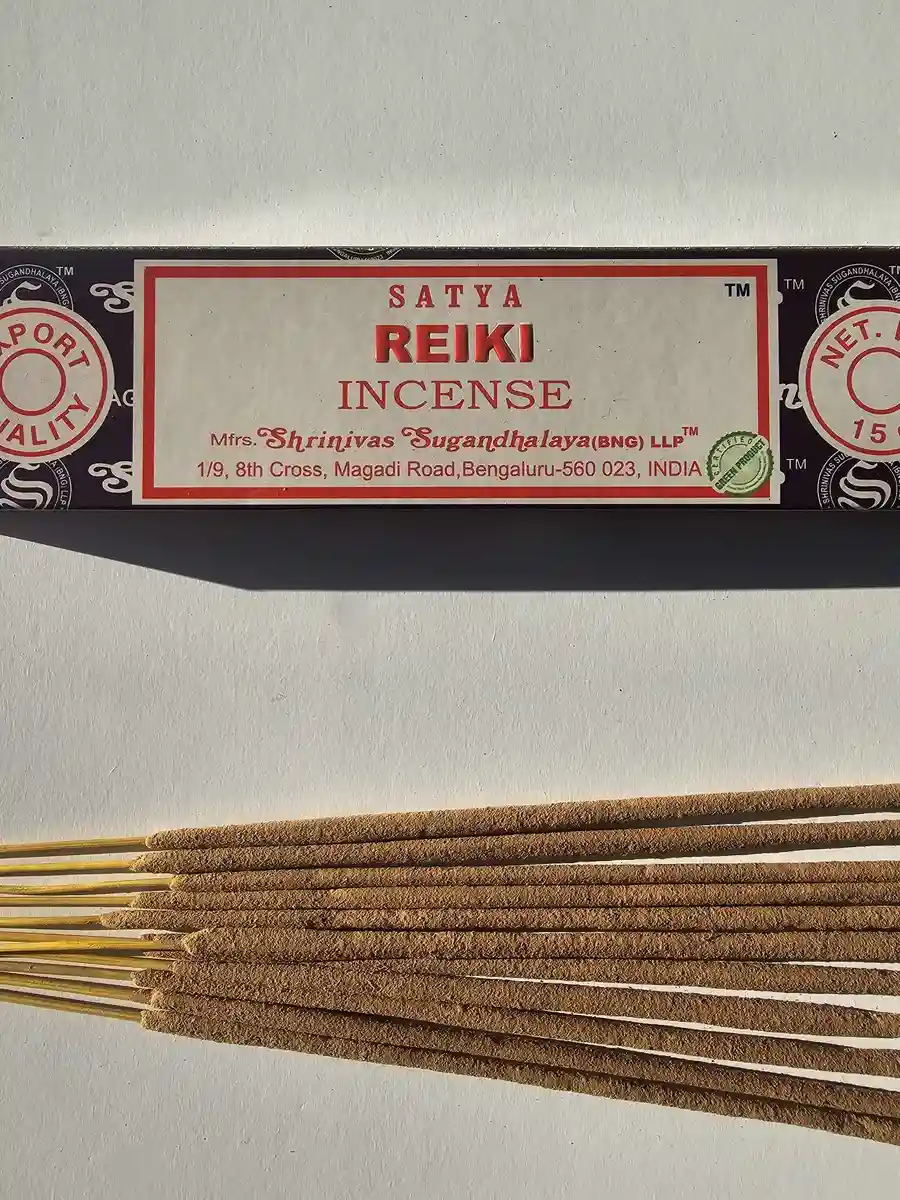 Ciemnofioletowe pudełko kadzideł Satya Reiki 15g umieszczone nad stosem jasnobrązowych patyczków na białym tle z wyraźnym cieniem.