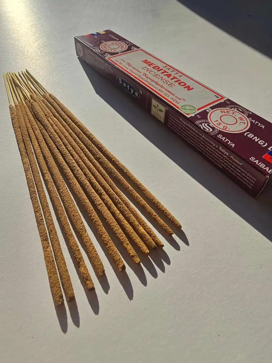 Bordowe pudełko kadzideł Satya Meditation 15g leżące nad stosem jasnobrązowych patyczków na białym tle z wyraźnym cieniem rzucanym w prawą stronę.