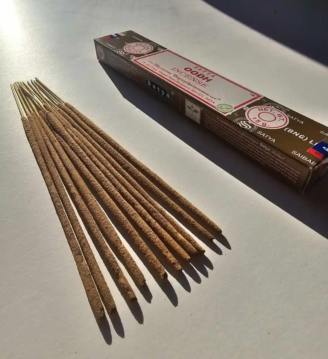 Ciemnobrązowe pudełko kadzideł Satya Oodh 15g leżące nad stosem jasnobrązowych patyczków na białym tle z wyraźnym cieniem rzucanym w prawą stronę.