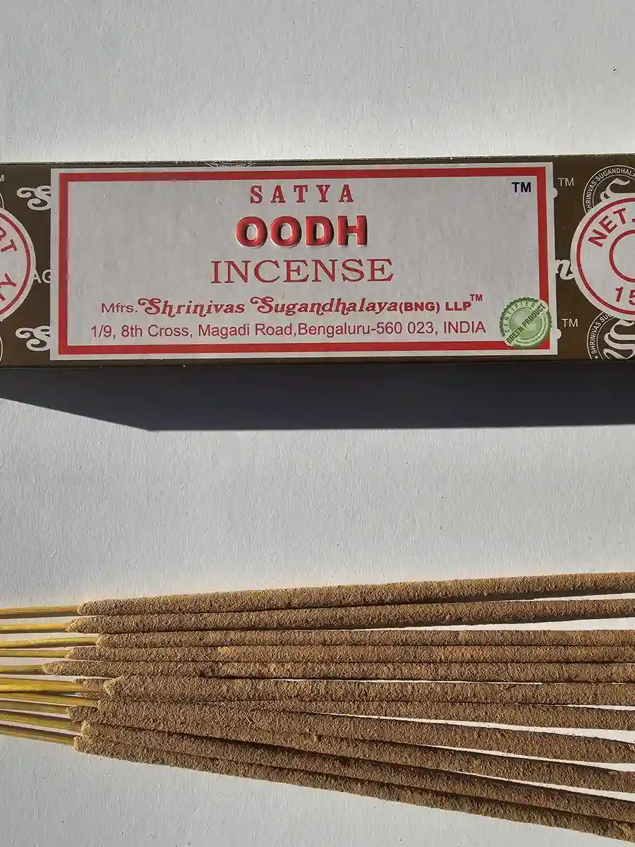 Ciemnobrązowe pudełko kadzideł Satya Oodh 15g leżące nad stosem jasnobrązowych patyczków na białym tle z wyraźnym cieniem rzucanym w prawą stronę.