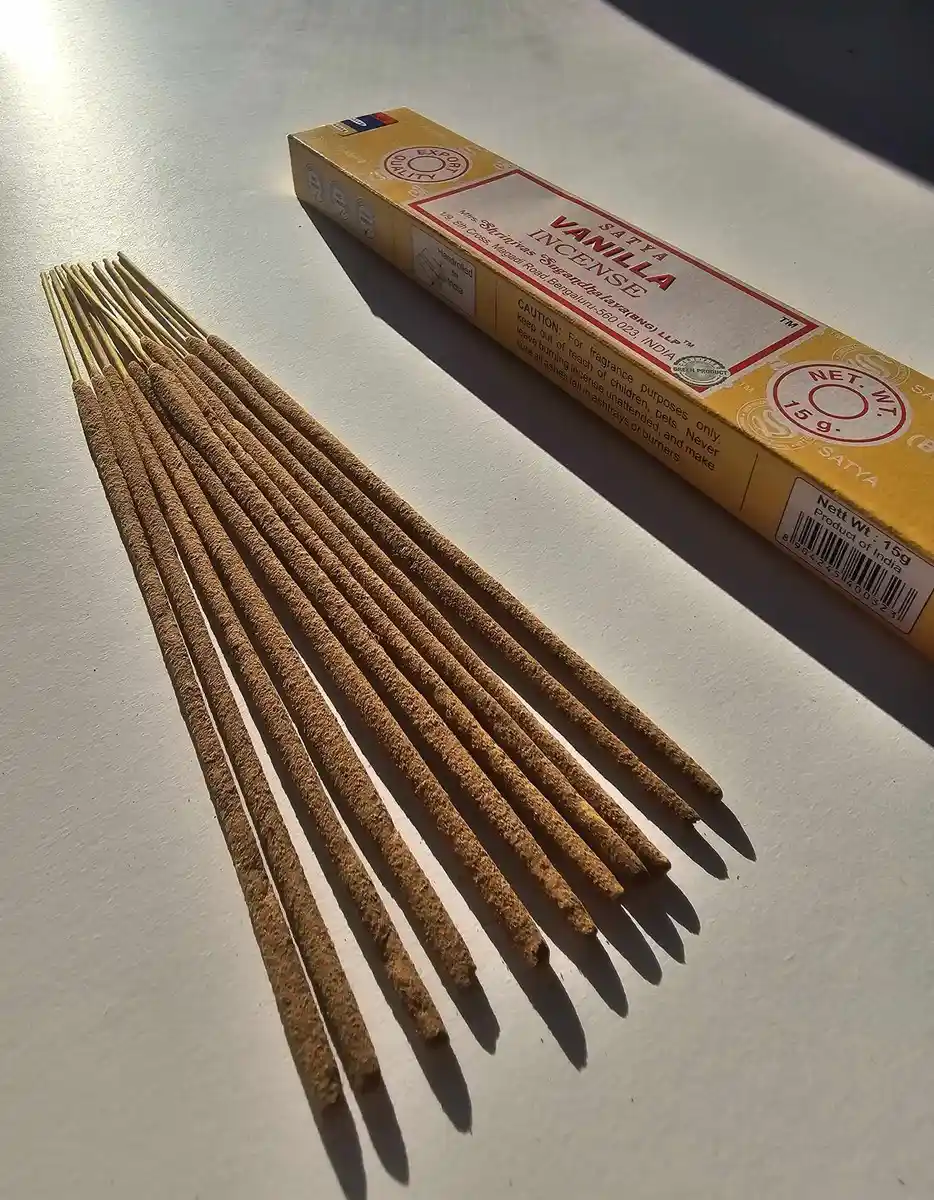 Jasnożółte pudełko kadzideł Satya Vanilla 15g leżące nad stosem jasnobrązowych patyczków na białym tle z wyraźnym cieniem rzucanym w prawą stronę.