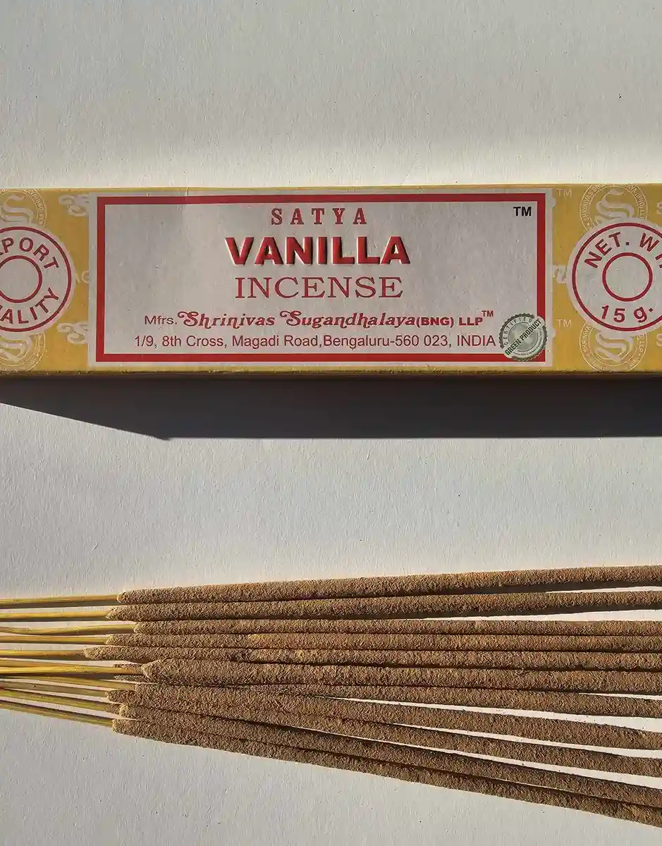 Jasnożółte pudełko kadzideł Satya Vanilla 15g leżące nad stosem jasnobrązowych patyczków na białym tle z wyraźnym cieniem rzucanym w prawą stronę.