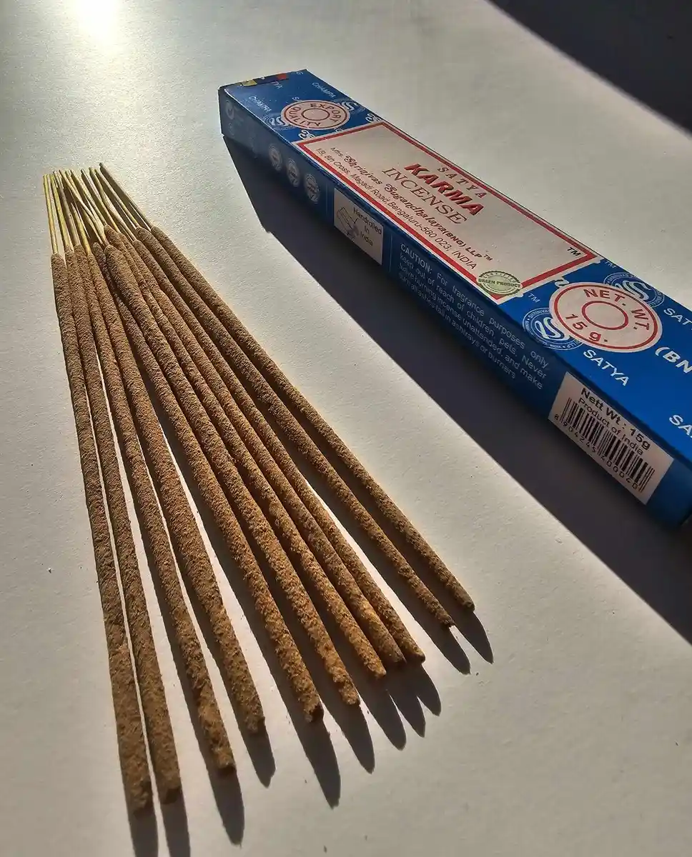 Niebieskie pudełko kadzideł Satya Karma 15g leżące nad grupą jasnobrązowych patyczków na białym tle z wyraźnym, poziomym cieniem.