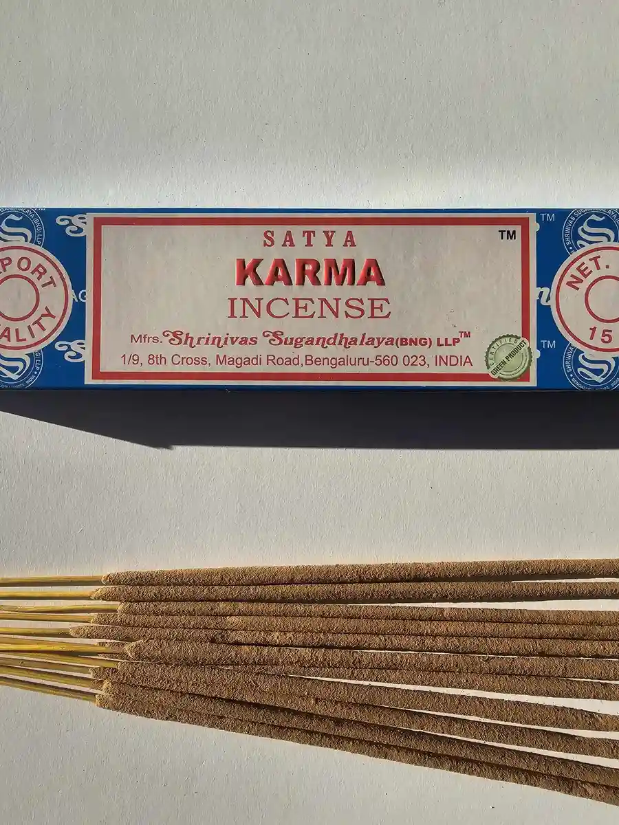 Niebieskie pudełko kadzideł Satya Karma 15g leżące nad grupą jasnobrązowych patyczków na białym tle z wyraźnym, poziomym cieniem.