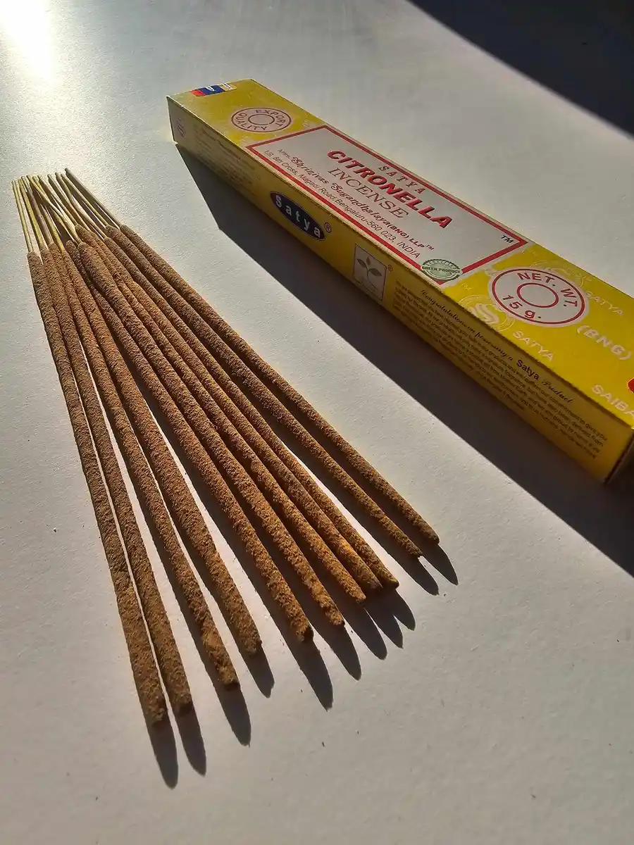 Jasnożółte pudełko kadzideł Satya Citronella 15g ułożone poziomo nad grupą jasnobrązowych patyczków na białym tle z wyraźnym cieniem.