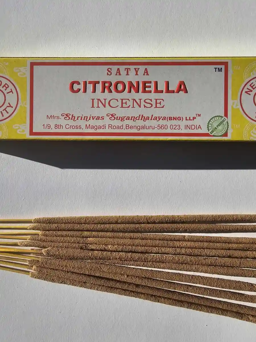 Jasnożółte pudełko kadzideł Satya Citronella 15g ułożone poziomo nad grupą jasnobrązowych patyczków na białym tle z wyraźnym cieniem.