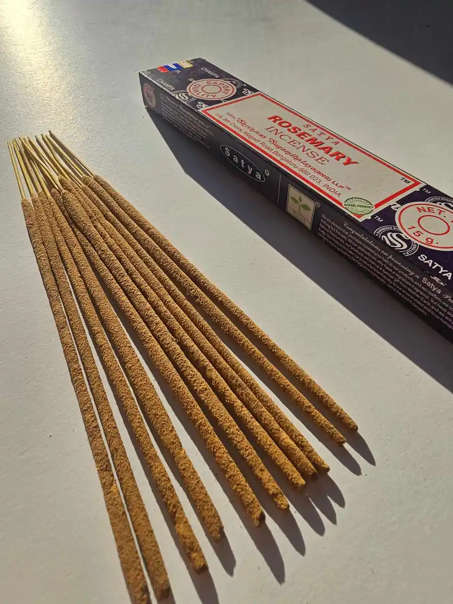 Ciemnofioletowe pudełko kadzideł Satya Rosemary 15g leżące poziomo nad stosem jasnobrązowych patyczków na białym tle z mocnym cieniem rzucanym w prawą stronę.