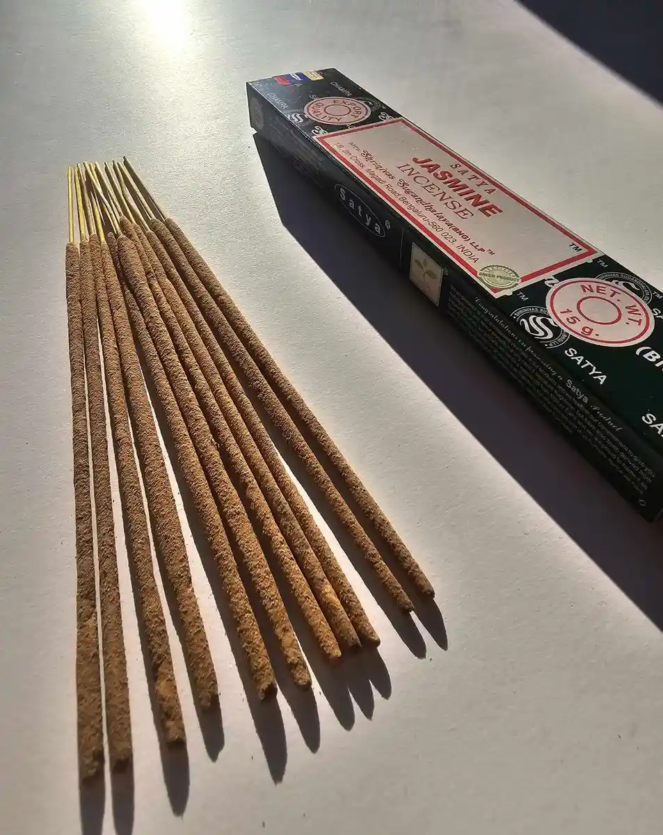 Ciemnozielone pudełko kadzideł Satya Jasmine 15g leżące nad stosem jasnobrązowych patyczków na białym tle z wyraźnym cieniem rzucanym w prawą stronę.