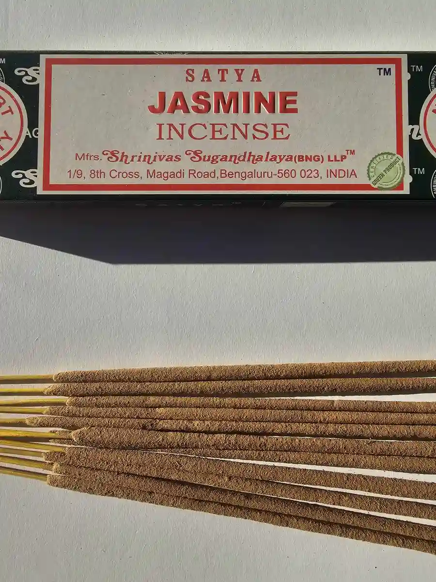 Ciemnozielone pudełko kadzideł Satya Jasmine 15g leżące nad stosem jasnobrązowych patyczków na białym tle z wyraźnym cieniem rzucanym w prawą stronę.