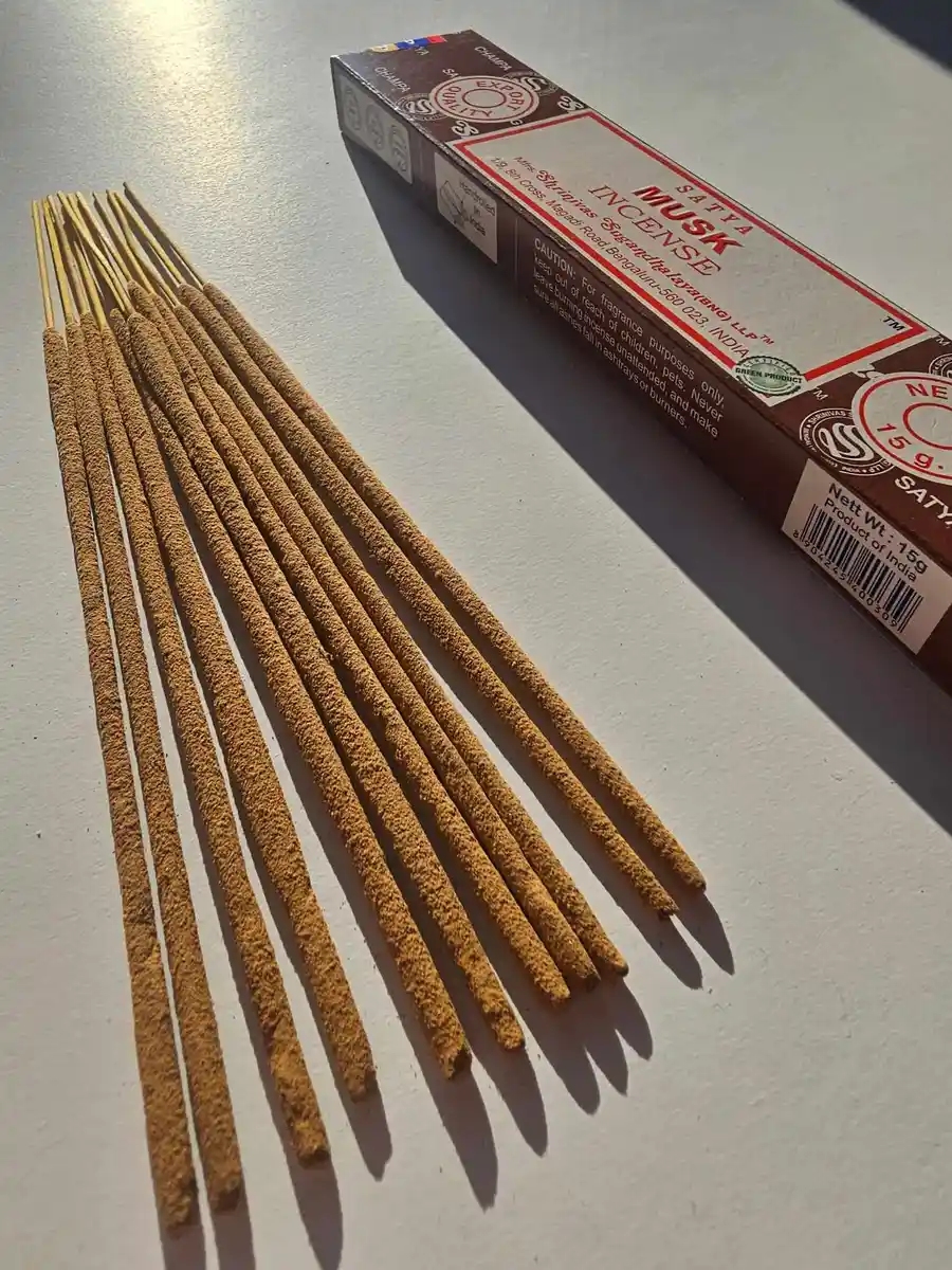Brązowe pudełko kadzideł Satya Musk 15g leżące nad stosem jasnobrązowych patyczków na białym tle z wyraźnym cieniem rzucanym w prawą stronę.