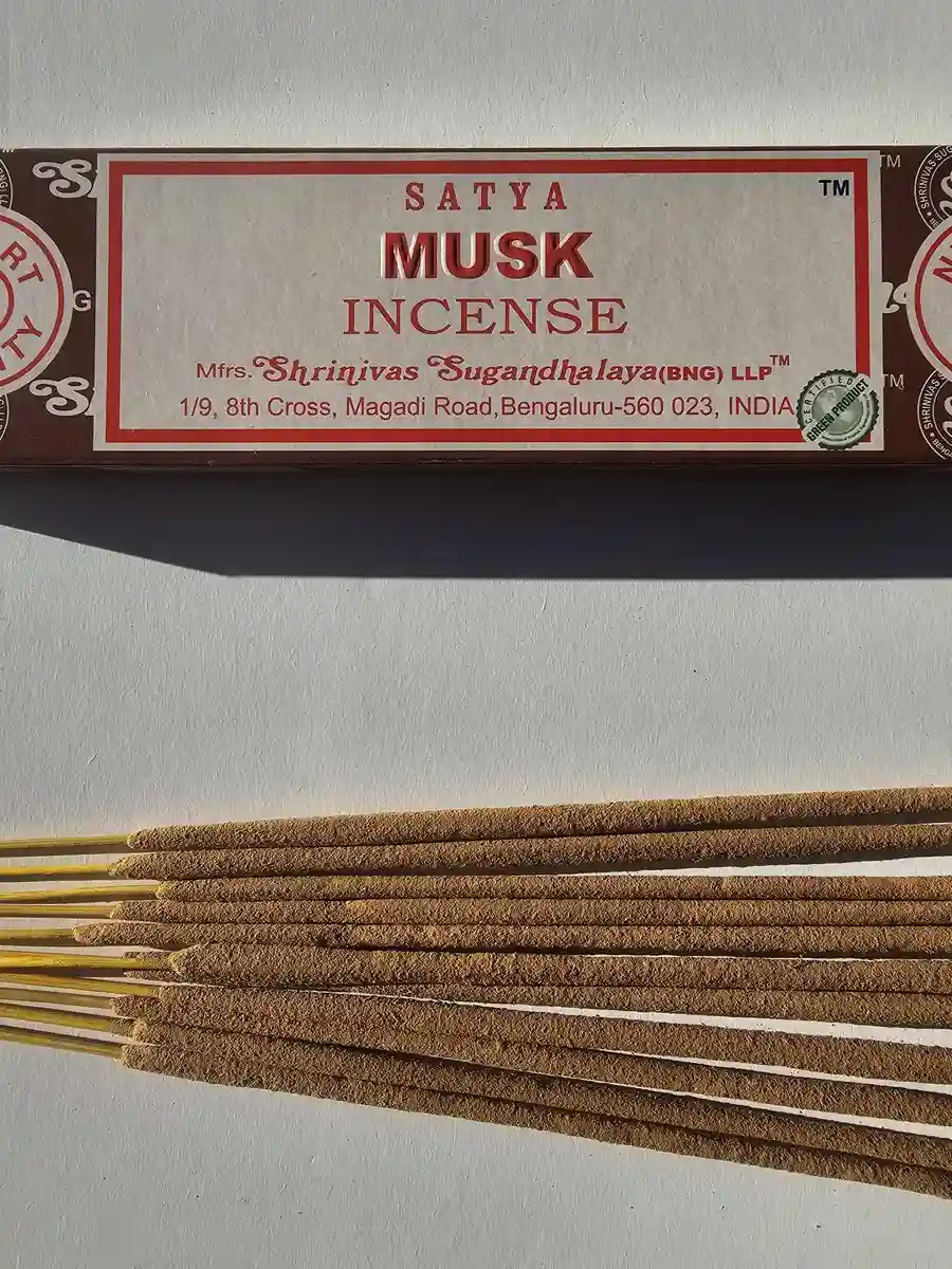 Brązowe pudełko kadzideł Satya Musk 15g leżące nad stosem jasnobrązowych patyczków na białym tle z wyraźnym cieniem rzucanym w prawą stronę.