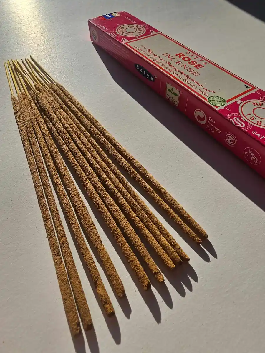 Różowe pudełko kadzideł Satya Rose 15g leżące poziomo nad stosem jasnobrązowych patyczków na białym tle z wyraźnym cieniem rzucanym w prawą stronę.