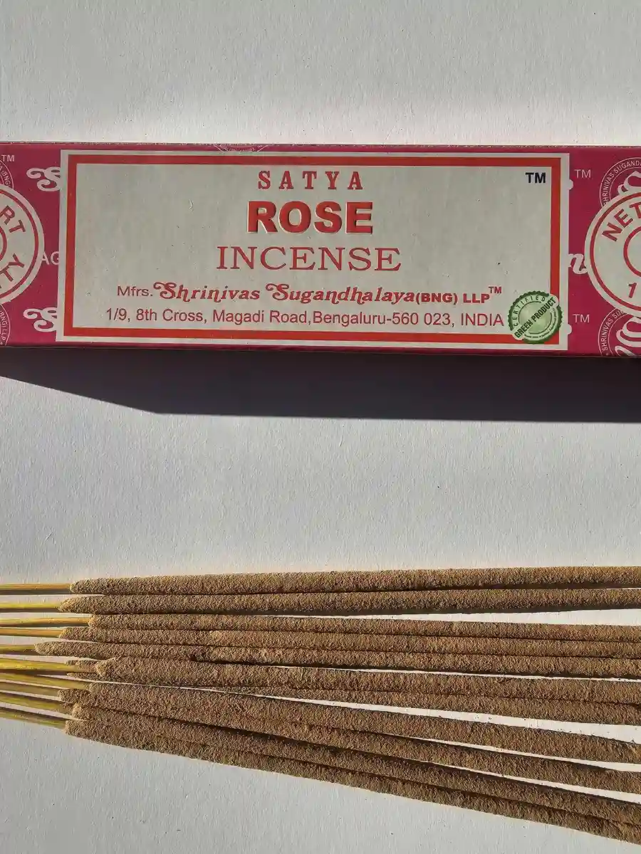 Różowe pudełko kadzideł Satya Rose 15g leżące poziomo nad stosem jasnobrązowych patyczków na białym tle z wyraźnym cieniem rzucanym w prawą stronę.