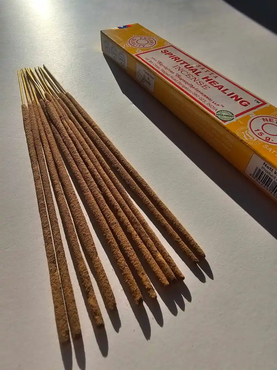 Jasnożółte pudełko kadzideł Satya Spiritual Healing 15g ułożone poziomo nad grupą jasnobrązowych patyczków na białym tle z wyraźnym, ostrym cieniem.
