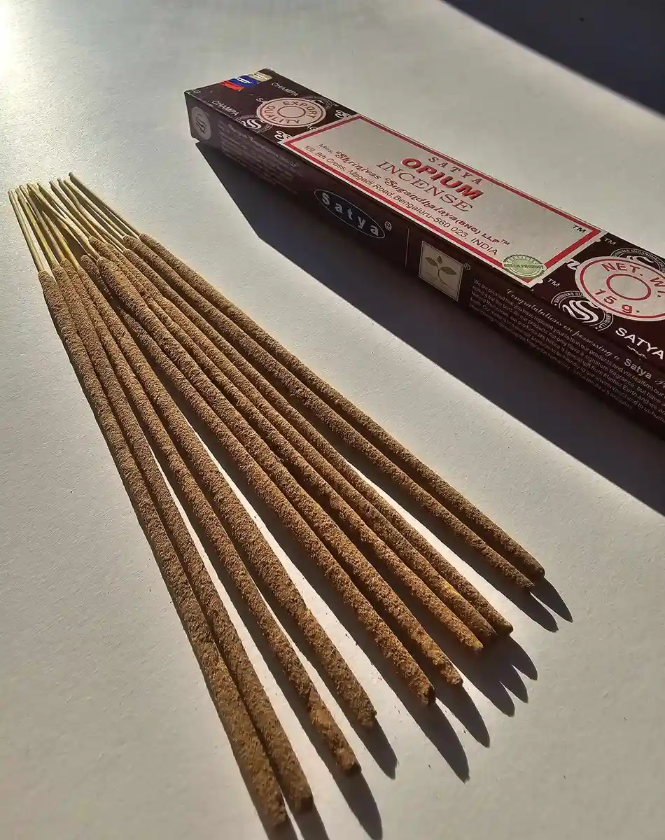 Ciemnobrązowe pudełko kadzideł Satya Opium 15g ułożone poziomo nad grupą jasnobrązowych patyczków na białym tle z wyraźnym, długim cieniem.