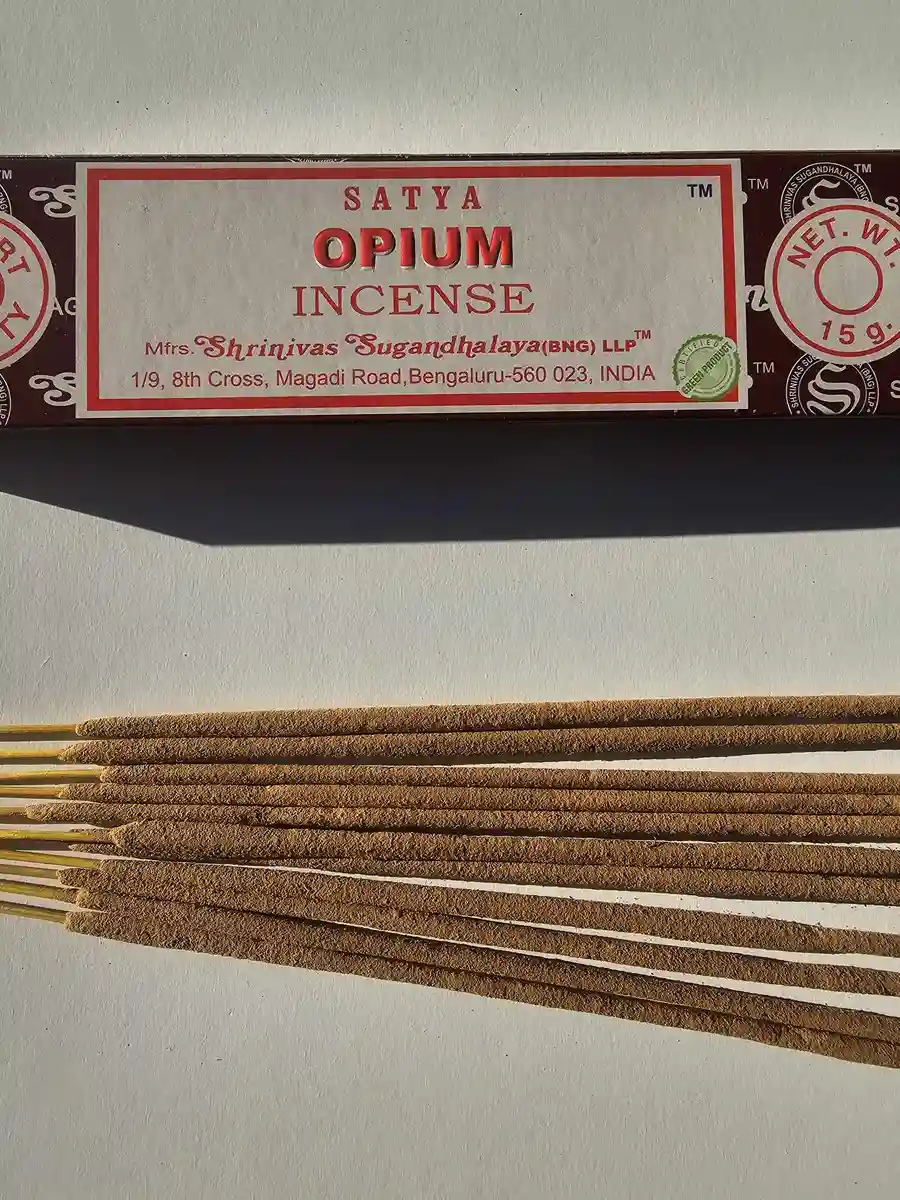 Ciemnobrązowe pudełko kadzideł Satya Opium 15g ułożone poziomo nad grupą jasnobrązowych patyczków na białym tle z wyraźnym, długim cieniem.