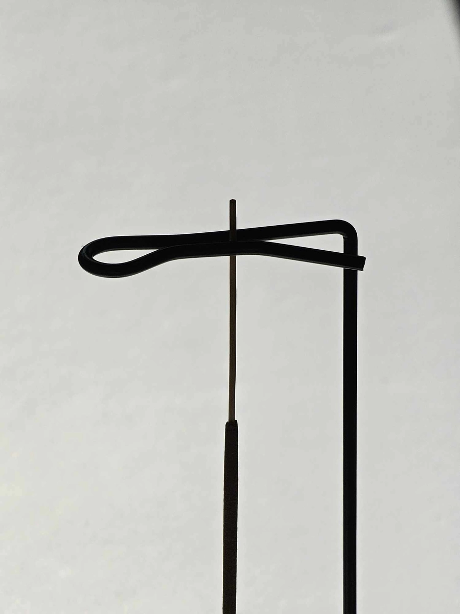 Elegancki metalowy uchwyt trzymający kadzidło patyczkowe do góry nogami nad drewnianą podstawką, prezentujący nowoczesny sposób palenia bez rozsypywania popiołu.