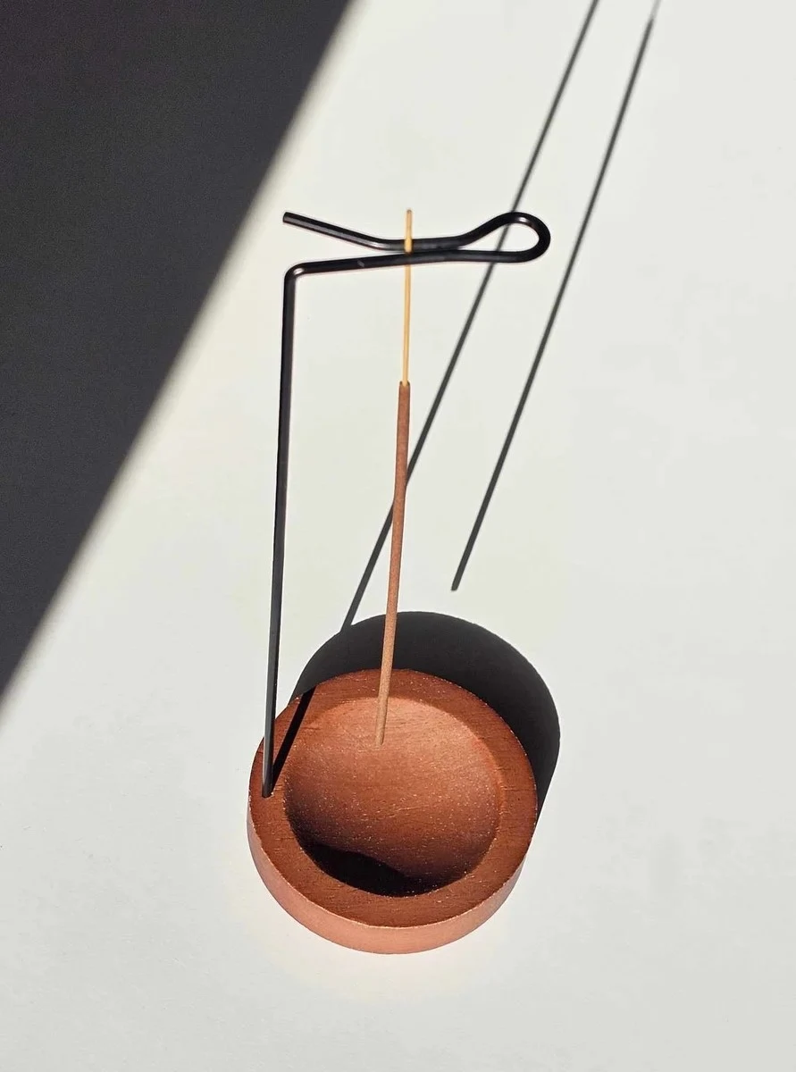 Elegancki metalowy uchwyt trzymający kadzidło patyczkowe do góry nogami nad drewnianą podstawką, prezentujący nowoczesny sposób palenia bez rozsypywania popiołu.